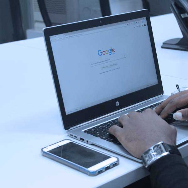 showing 2 male hands typing on a notebook computer keyboard. The computers screen is showing the home page for Google, ready to type in a search. The notebook computer is sitting on a large white desk and there is a mobile phone next to the laptop.